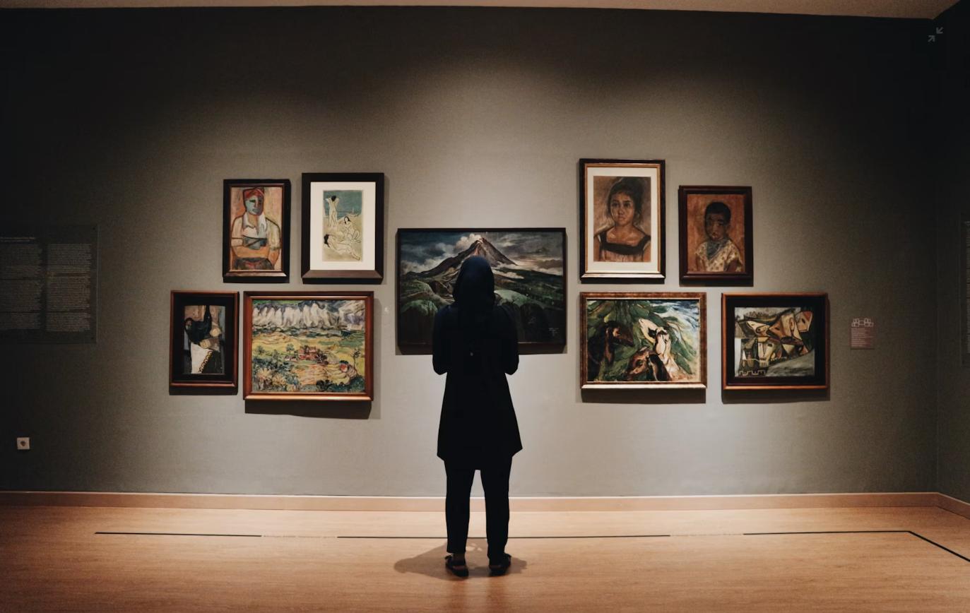 A visitor viewing a wall of framed paintings inside a quiet gallery space